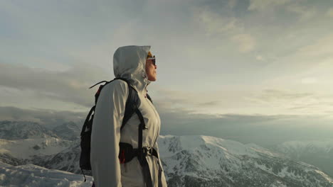 hiker stands looking out over dramatic snow capped ridges in soft morning light, orbit slow motion, upward angle