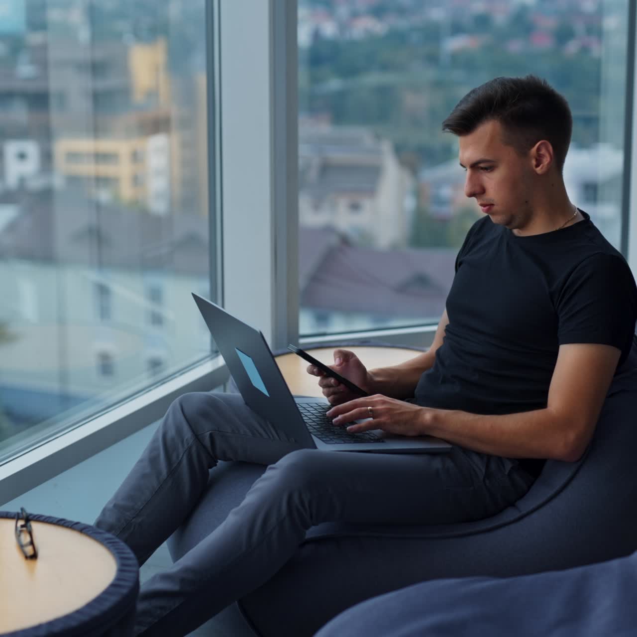 Young man sitting in bean-bag chair with laptop on his knees. Businessman looking at phone in his hands. Cityscape at backdrop in blur