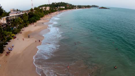 aerial dolly in of people having fun in turquoise sea and sand shore near hotels and forest in mirissa beach, sri lanka