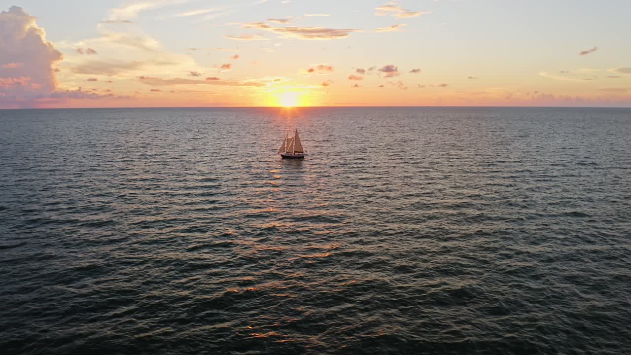 A lone sailboat drifts across rippling Gulf waters beneath a glowing sunset, framed by warm skies and clouds that create a serene evening seascape