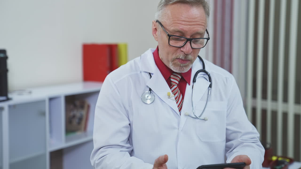 Senior doctor with a phone in medical center. Grey haired therapist in medical white coat with stethoscope giving consultation through the mobile phone in clinic.