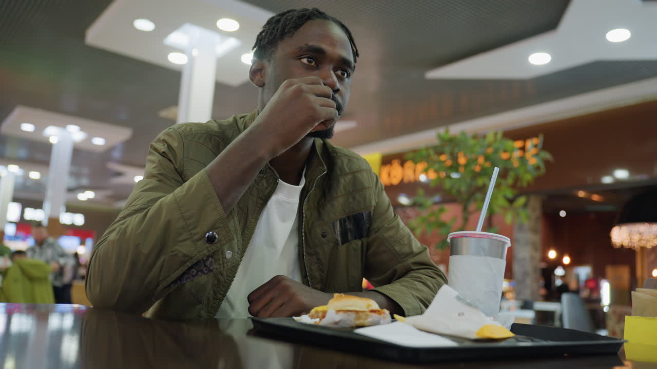 Student in casual jacket sits at food court table eating meal with drink beside tray containing burger and fries while looking ahead indoors under bright ceiling lights and blurred background activity