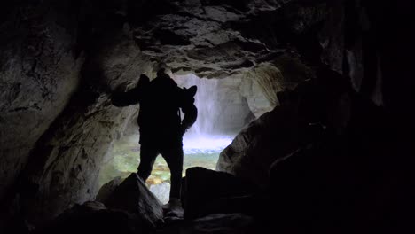 Man-exploring-cave,-heading-towards-a-small-waterfall-at-the-end-of-the-tunnel