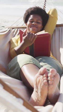 feliz mujer afroamericana leyendo y acostada en una hamaca en una playa soleada