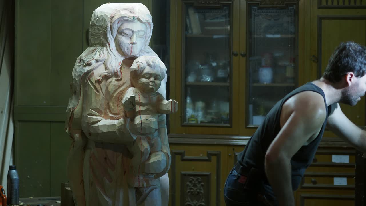A sculptor leaves his workbench to select a new tool, while a partially carved wooden Madonna and Child statue stands in front of vintage workshop cabinets