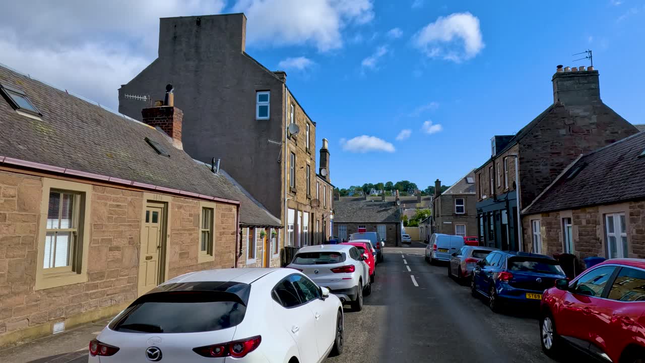 A vehicle travels slowly along a narrow residential street lined with parked cars and historic stone buildings under bright daylight and clear blue skies