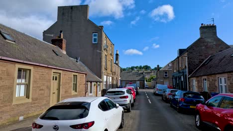 a vehicle travels slowly along a narrow residential street lined with parked cars and historic stone buildings under bright daylight and clear blue skies