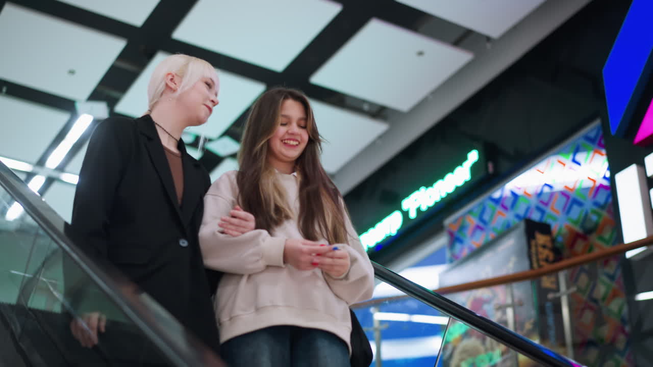 Close friends descend on escalator indoors laughing and discussing while holding arms, captured from low angle with modern ceiling panels and bright mall lights
