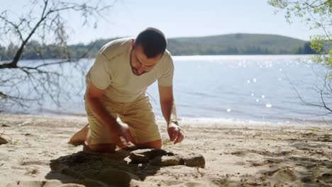 Mann-Macht-Ein-Feuer-Auf-Sand-Am-Strand-Mit-Ruhigem-Meer-Im-Hintergrund