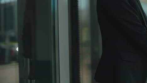 close up of professional woman with handbag opening glass door stepping out of building, focus on hand gripping metal handle, reflections visible on door, modern urban setting with sleek architecture