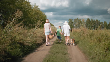 back view of family walking on dirt road surrounded by lush greenery during retreat, carrying picnic basket while aunty holds umbrella and dog on leash, under partly cloudy sky