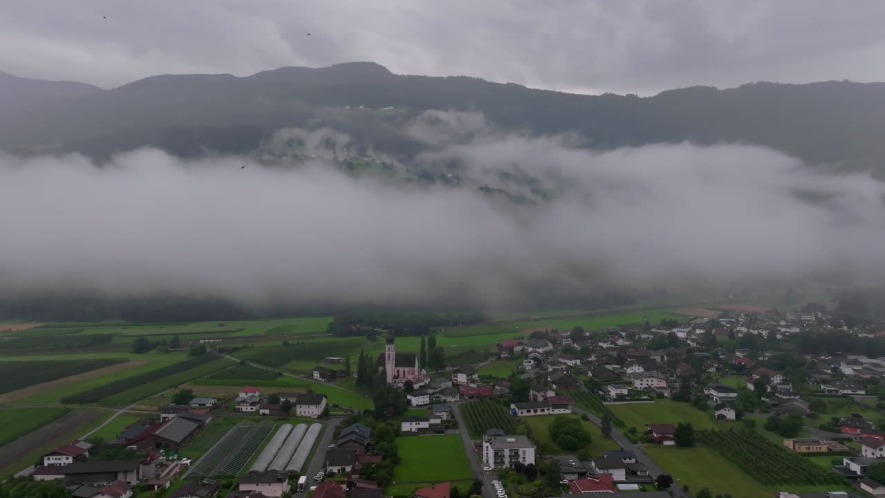 Letecký pohled na vesnici v rakouských Alpách s kostelem v popředí a mlhou valící se přes hory