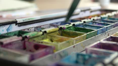 close-up of a painting brush being quickly dipped into green watercolor paint and then put on a wooden table next to the palette