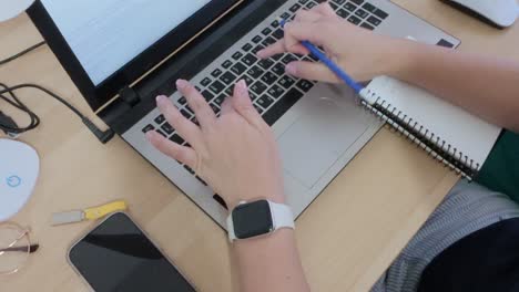 Person working remotely, writing notes in a spiral notebook while using a laptop