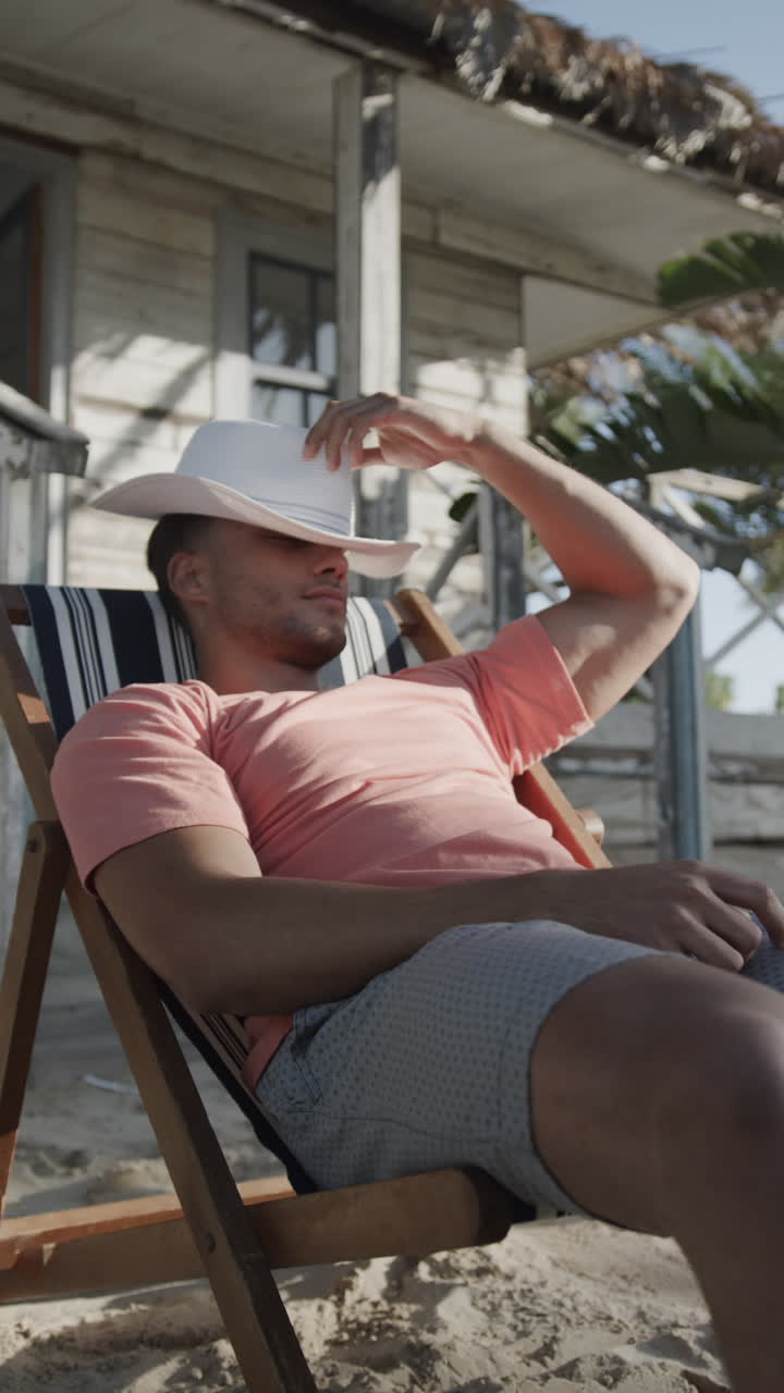 Vertical video of happy latino man with hat on face sitting in sun by beach house, slow motion