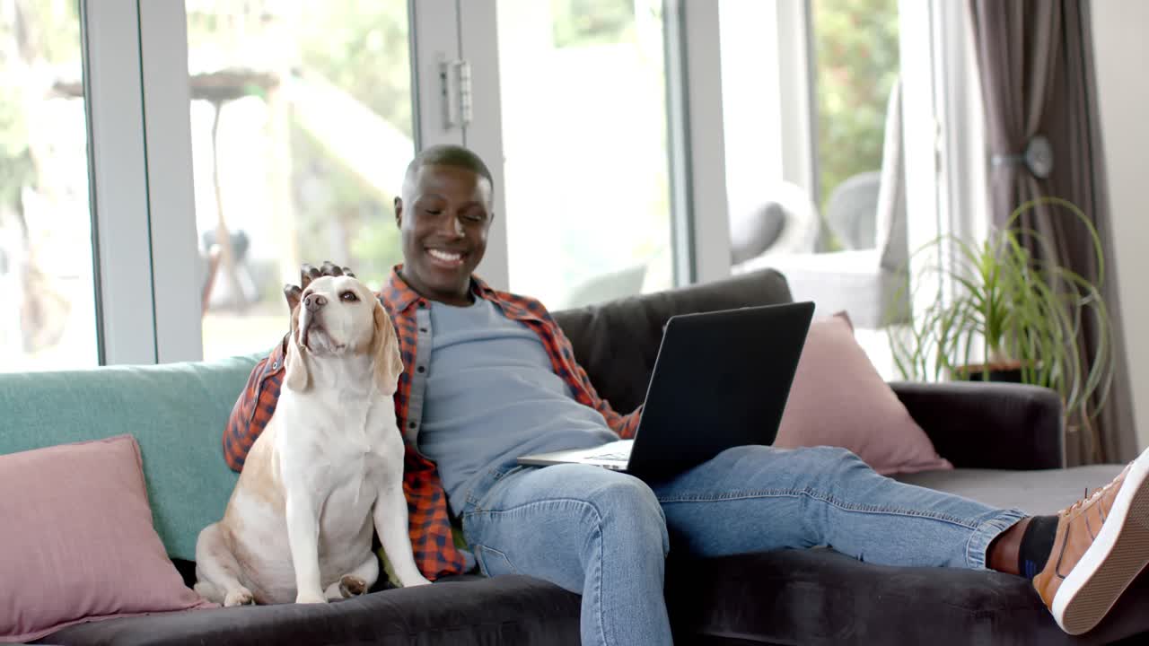 Happy african american man sitting on sofa using laptop at home, with his pet dog, slow motion