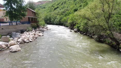 Drone-view-in-Albania-flying-over-a-moving-rapids-river-between-green-mountains-on-a-sunny-day-and-a-brick-house