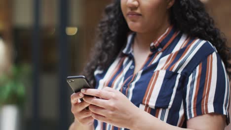 una mujer de negocios biracial sonriente de pie en el pasillo usando un teléfono inteligente en una oficina moderna