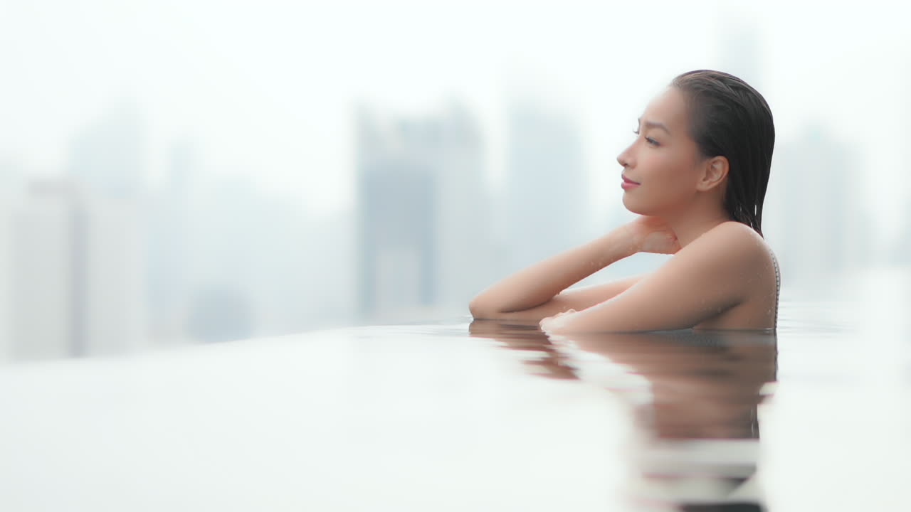 Resting on the edge of an infinity pool a young woman looks over the urban skyline in the distance. Copy space