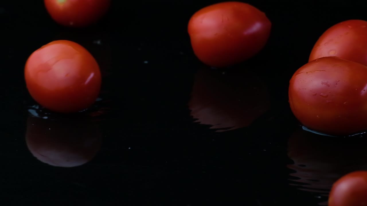 Cherry tomatoes sit in shallow water reflecting red juiciness in the water with black background