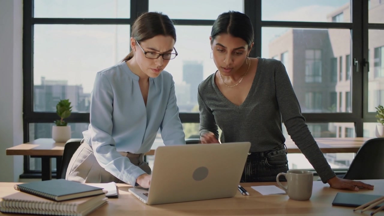 Reunión comenzando, dos compañeras vestidas de negocios colaborando con un portátil en un escritorio de oficina.
