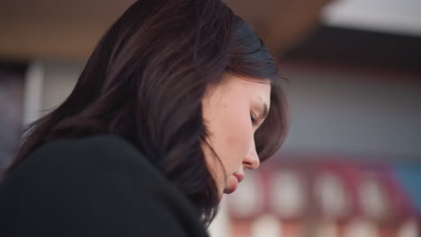 close-up profile of young woman with soft features and pink lips, looking down, focused, with blurred outdoor background featuring architectural structures and outdoor seating