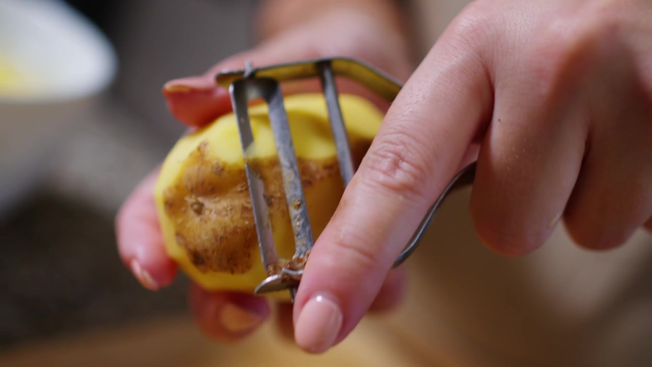 Primer plano en cámara lenta de las manos de un modelo usando un pelador de patatas para pelar una patata, centrándose en el proceso de pelado en un entorno de cocina