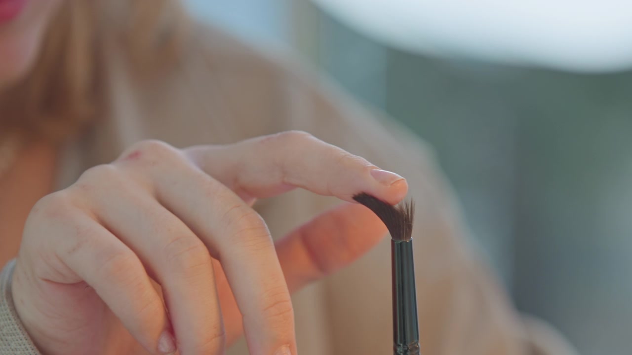 Close up of woman hands gently holding and rotating eyeshadow brush demonstrating its shape. soft lighting highlights texture of both hand skin and bristles against blurred background