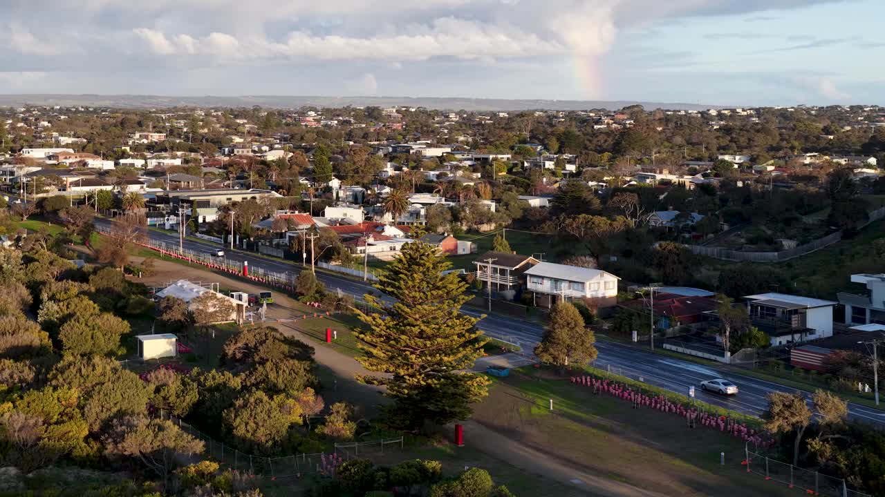 Drone glides above coastal road and shoreline, revealing seaside town, trees, and calm bay waters