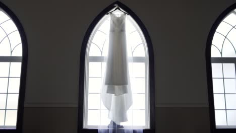 wide vista of a bride's beautiful wedding dress hanging from a large window sill in an old church building - slow push in slider shot