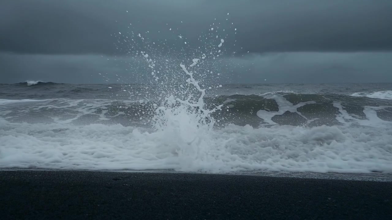 灰色の空の下、中央で波が砕け高いしぶきを上げ、押し寄せるうねりが暗い砂浜に打ち寄せている。