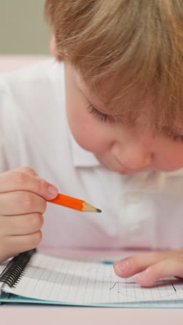 preschooler draws picture with pencil in notebook sitting at desk. blond boy touches chin thoughtfully. child develops artistic skills closeup