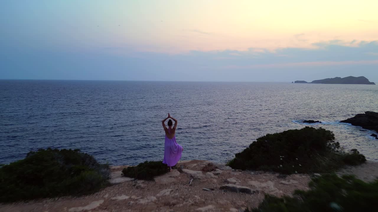 Woman meditating in yoga position on a cliff overlooking the sea at sunset in Ibiza. Magic aerial view flight overflight flyover drone