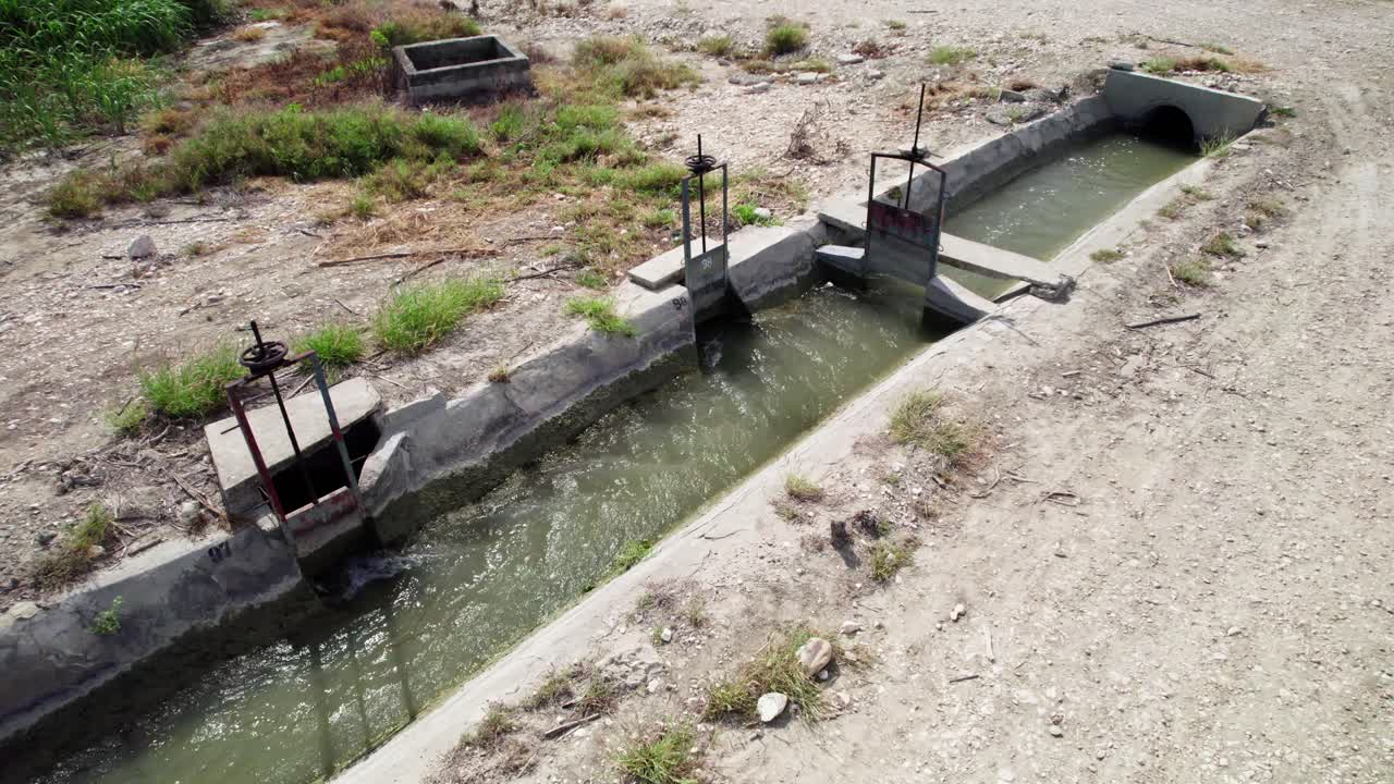 Toma aérea en semiórbita alrededor de un canal de riego de hormigón que transporta agua a través de tierras agrícolas en Barahona durante un día claro y soleado