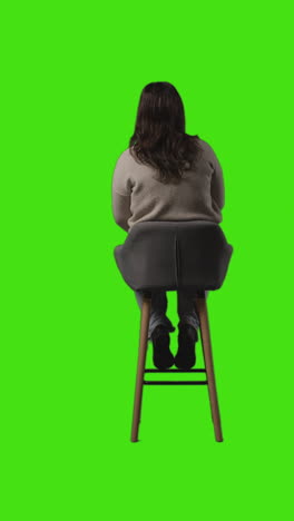 녹색 스크린 1에 대항하여 좌석에 앉아 기다리고있는 캐주얼 옷을 입은 젊은 여성의 수직 뒷면 전체 길이