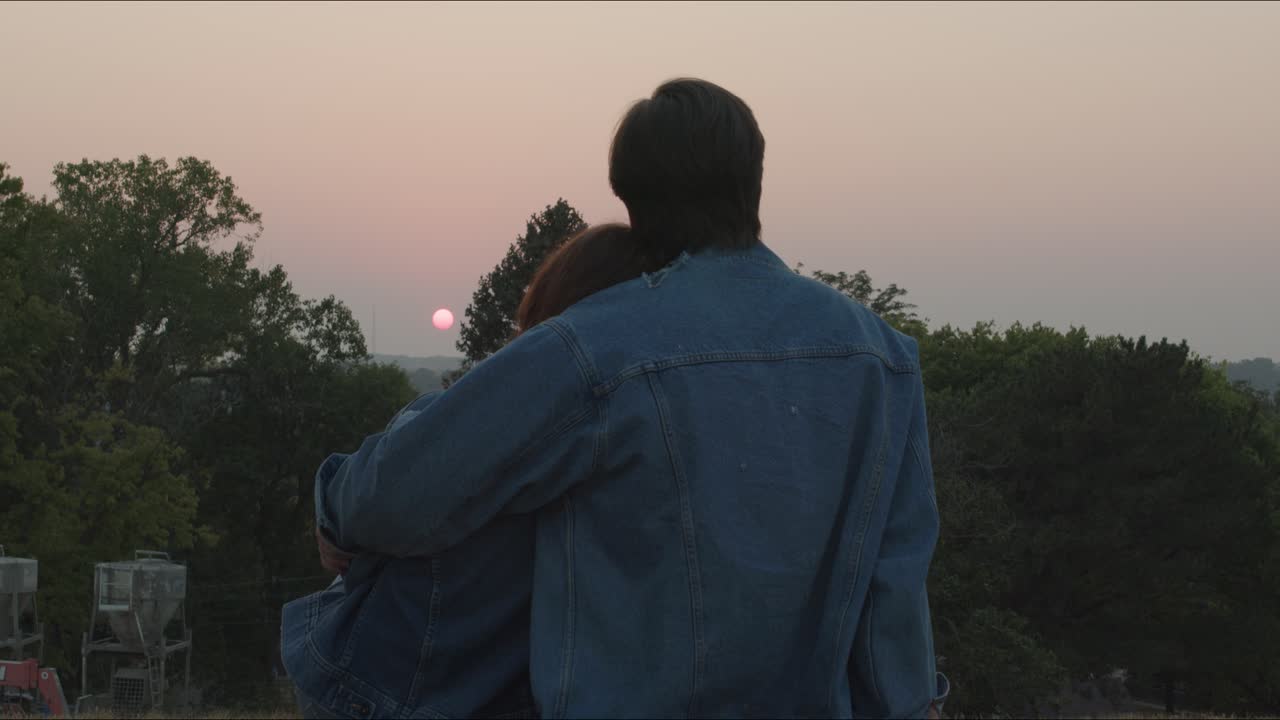 Man hugs woman while they sit in a park watching the sunset