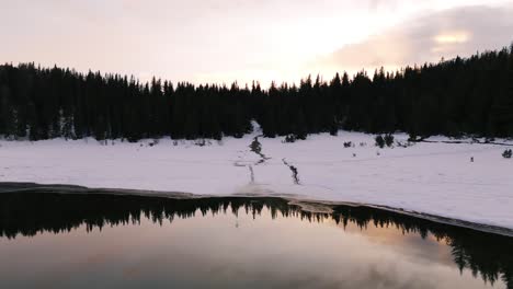 reverse aerial reveals winter sunrise and forest reflection in clear alpine lake