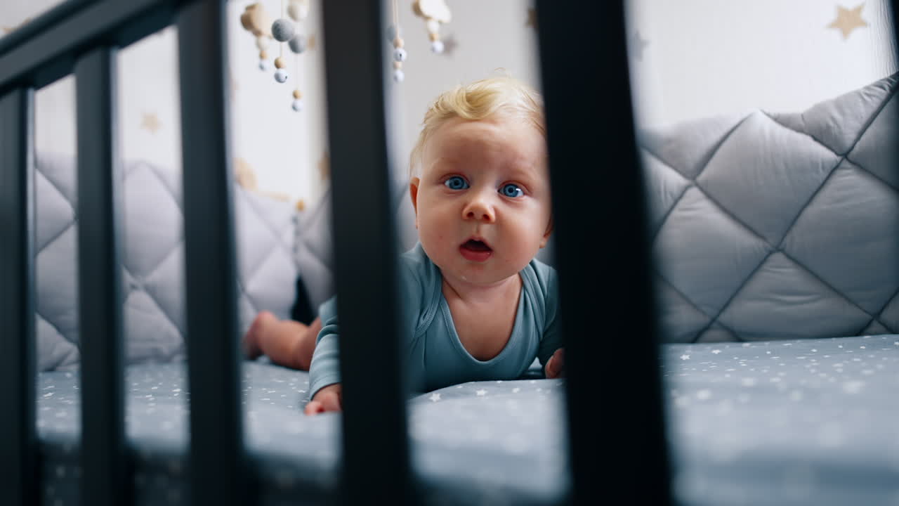 Looking into the crib with little baby lying inside. Beautiful infant kid looking at camera with interest.