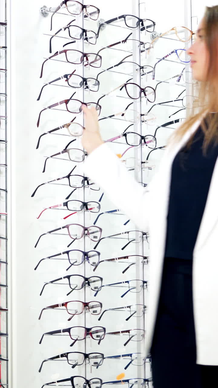 Girl in new glasses looking at camera at optics. Attractive young woman trying on fashionable eyeglasses indoors. Happy buyer choosing spectacles. Vertical video