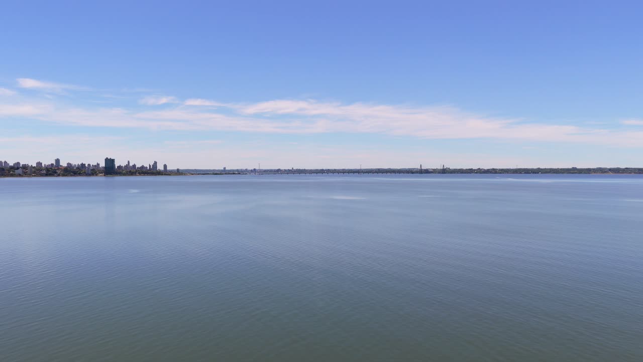 Aerial over Paraná River at Miguel Lanús, Posadas: curved walkway on green bank, water expanse, and distant skyline under blue sky in Misiones, Argentina