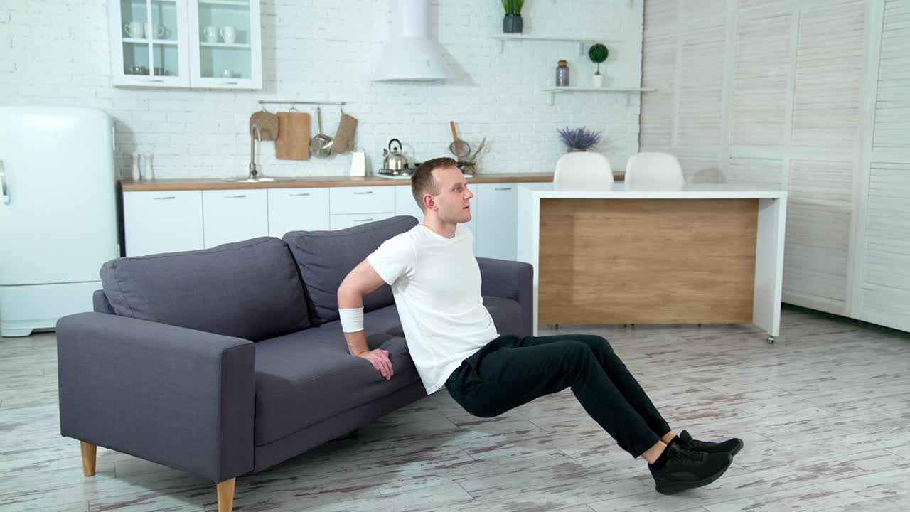 Young man exercising at home. Fellow in white t-shirt and black pants doing exercises near the sofa during lockdown. Slow motion.