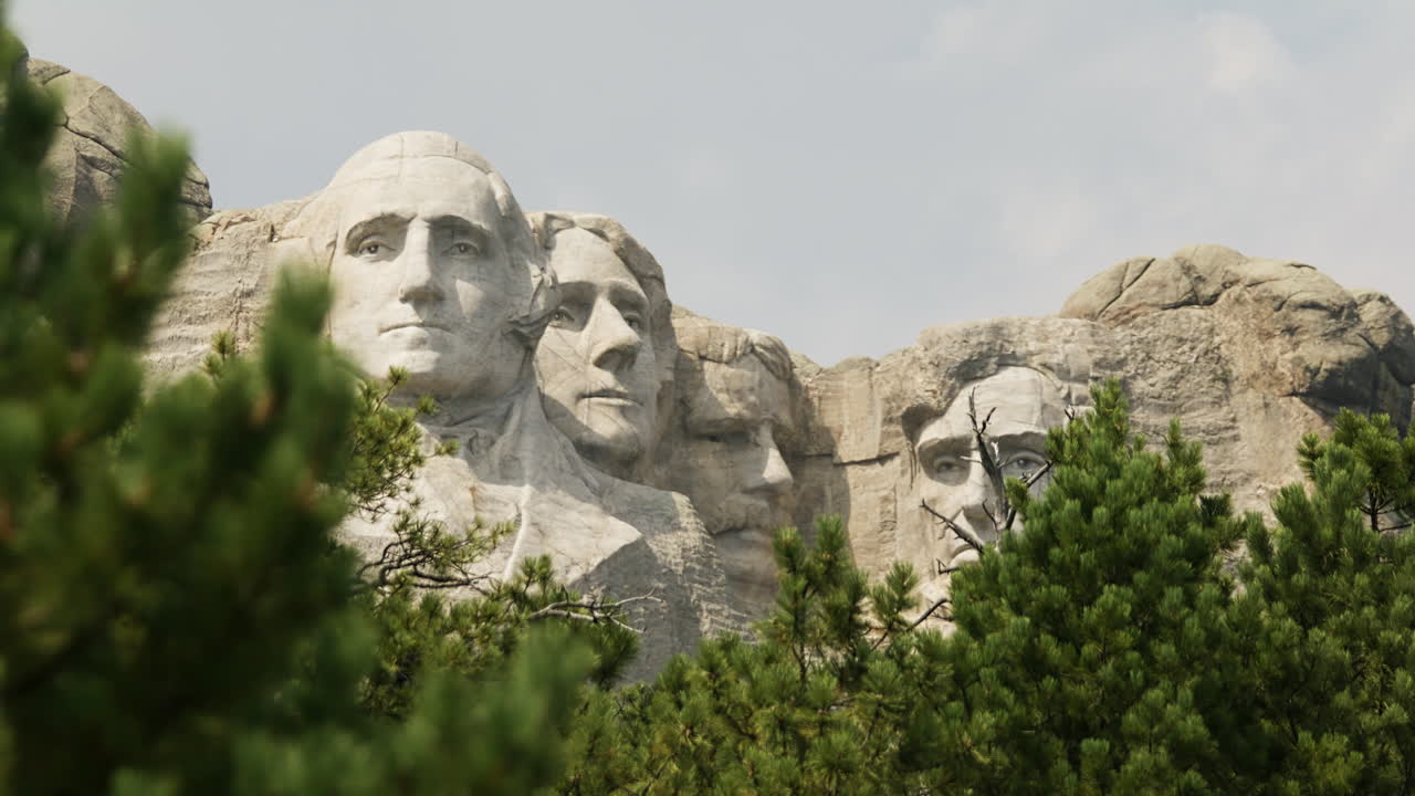 Inquadratura fissa del Mount Rushmore National Memorial, con pini in primo piano; cielo luminoso.