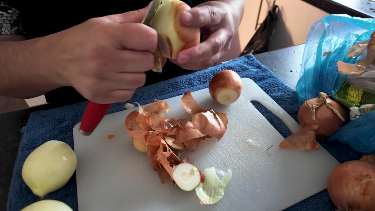 Close up of hands peeling an onion with a knife on a cutting board, preparing ingredients for a meal
