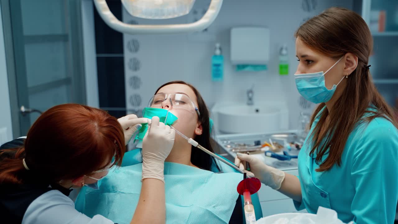 Young woman treats her sick tooth in clinic. Female dentist and assistant treating client's teeth in a modern dental office.