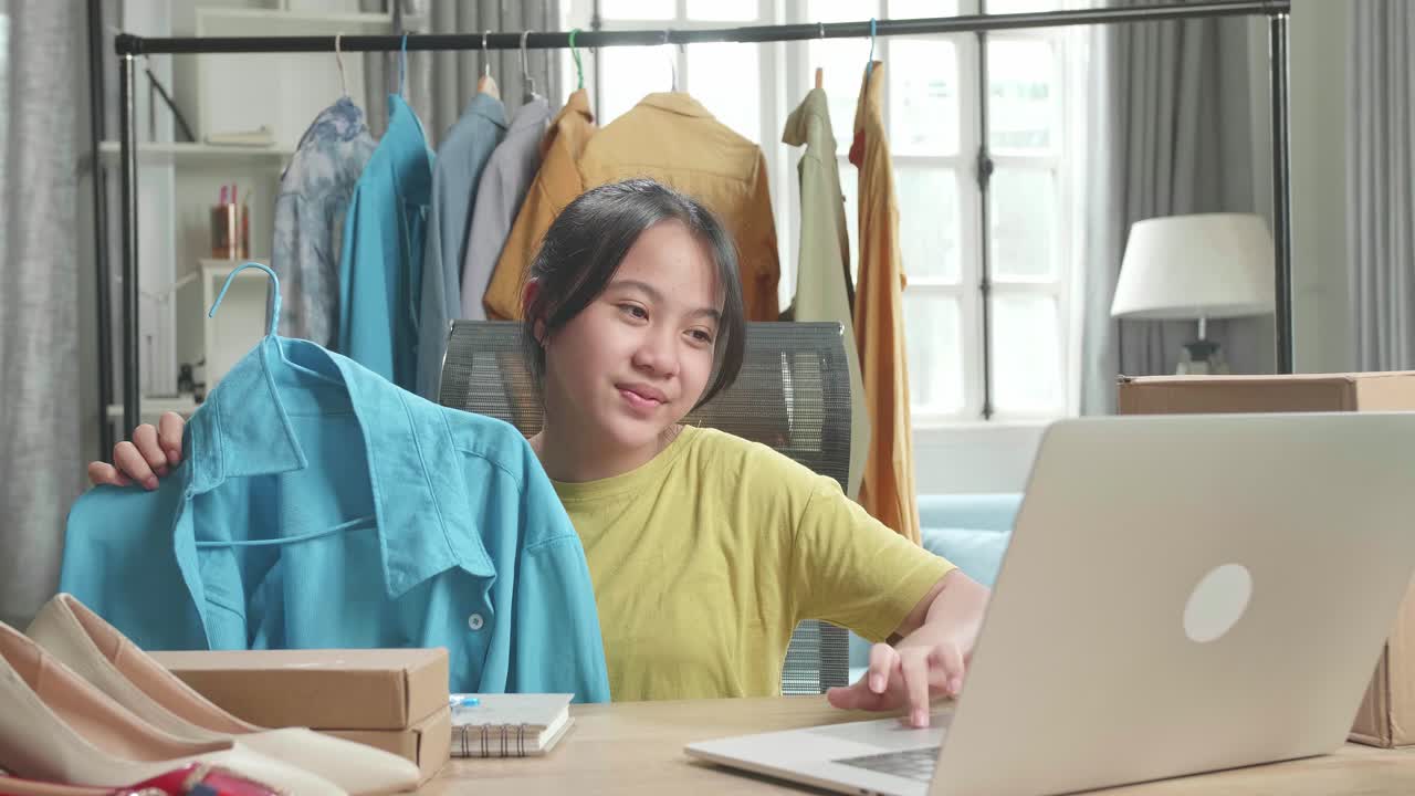 Gadis Muda Asia Penjual Online Merekomendasikan Kemeja Sambil Menggunakan Komputer untuk Menjual Pakaian di Rumah