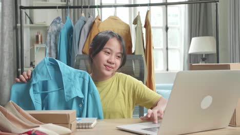asian young girl online seller recommending a shirt while using computer for selling clothes at home
