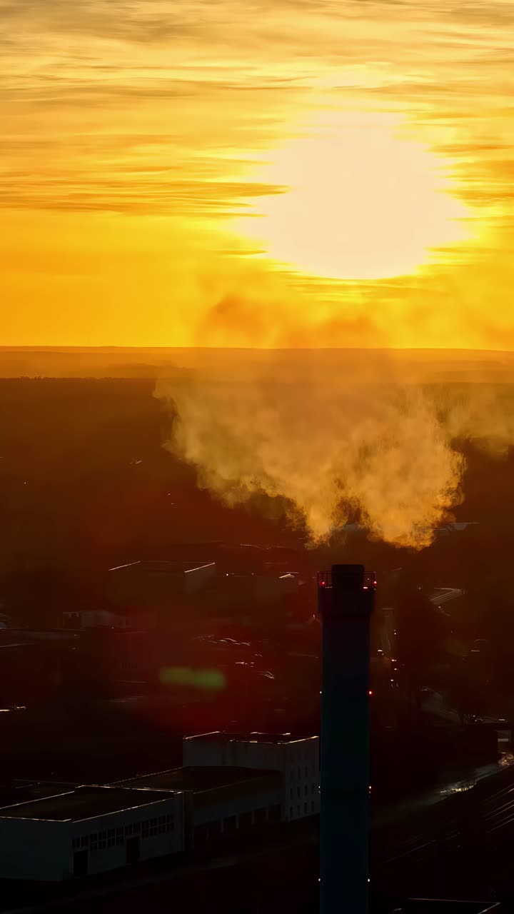 Gases rise from an industrial chimney against a golden sky at sunset. Vertical shot.