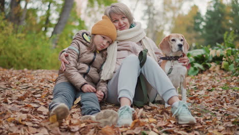 mother sits with daughter leaning against her shoulder while dog rests beside them on dry autumn leaves in quiet forest, showing warmth, comfort, family bonding, and gentle moments in nature together