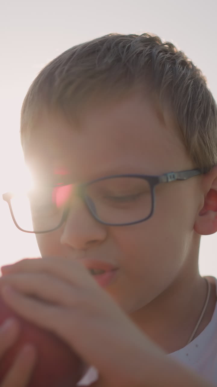 Close-up shot of a young boy wearing a white shirt and glasses, happily eating a red apple in a sunlit field. His expression is content, capturing a joyful moment of simplicity and nature