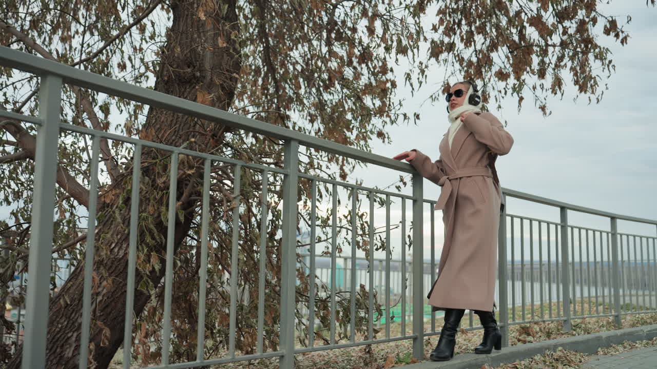 Una donna in trench posa su un belvedere cittadino, con cuffie e una borsa in pelle, tra foglie autunnali e un cielo coperto. Un passo elegante e una calma sicurezza creano un ritratto urbano cinematografico.